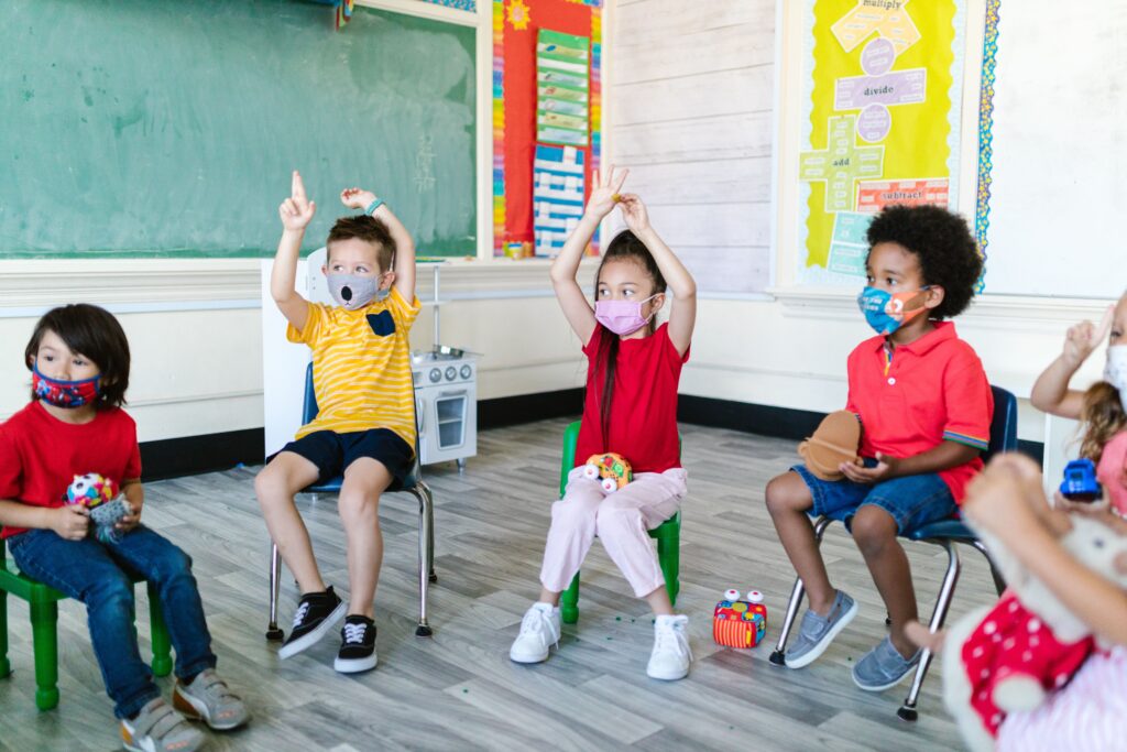 Children in masks actively participate in a lively classroom environment, showing excitement and engagement.
