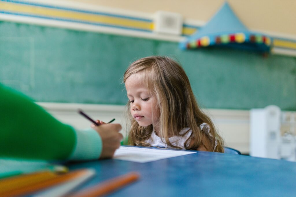A young girl with long hair concentrates on her drawing in a colorful classroom environment.