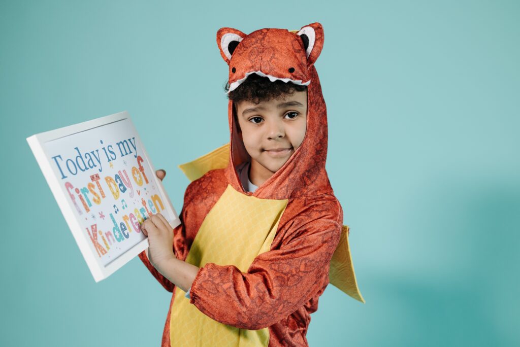 A young child in a dinosaur costume proudly holds a kindergarten first day sign.