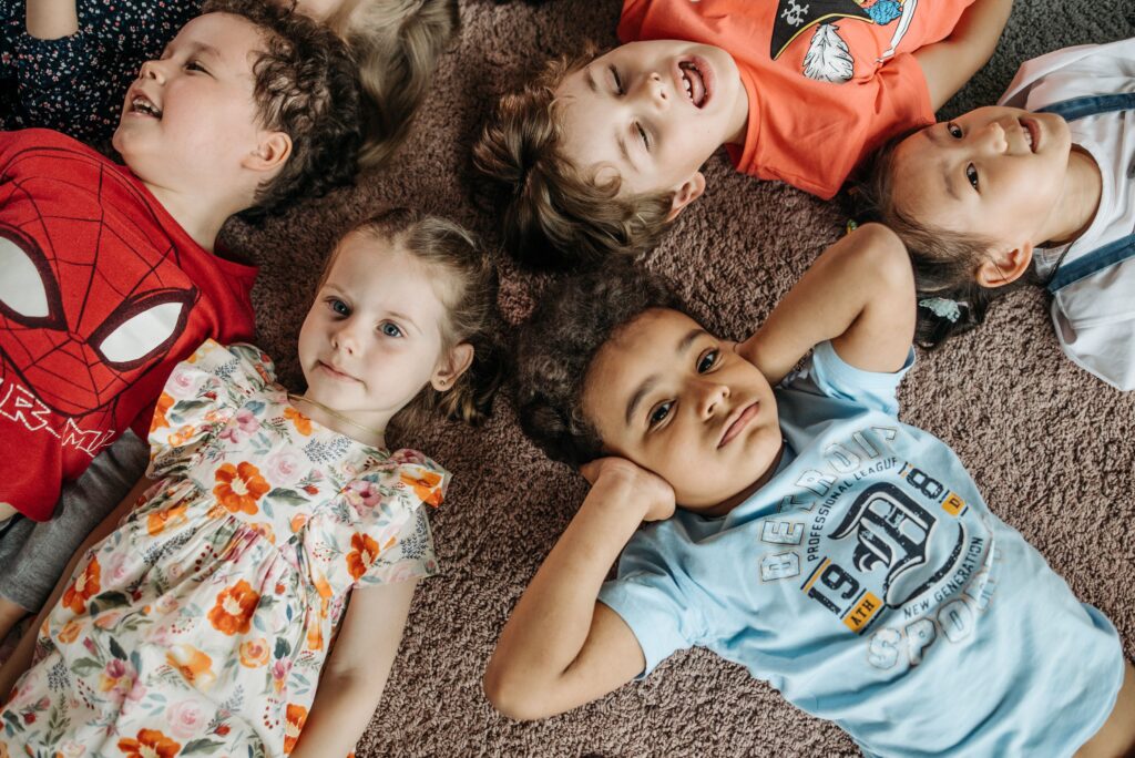 A cheerful group of diverse children lying playfully on a carpet indoors.