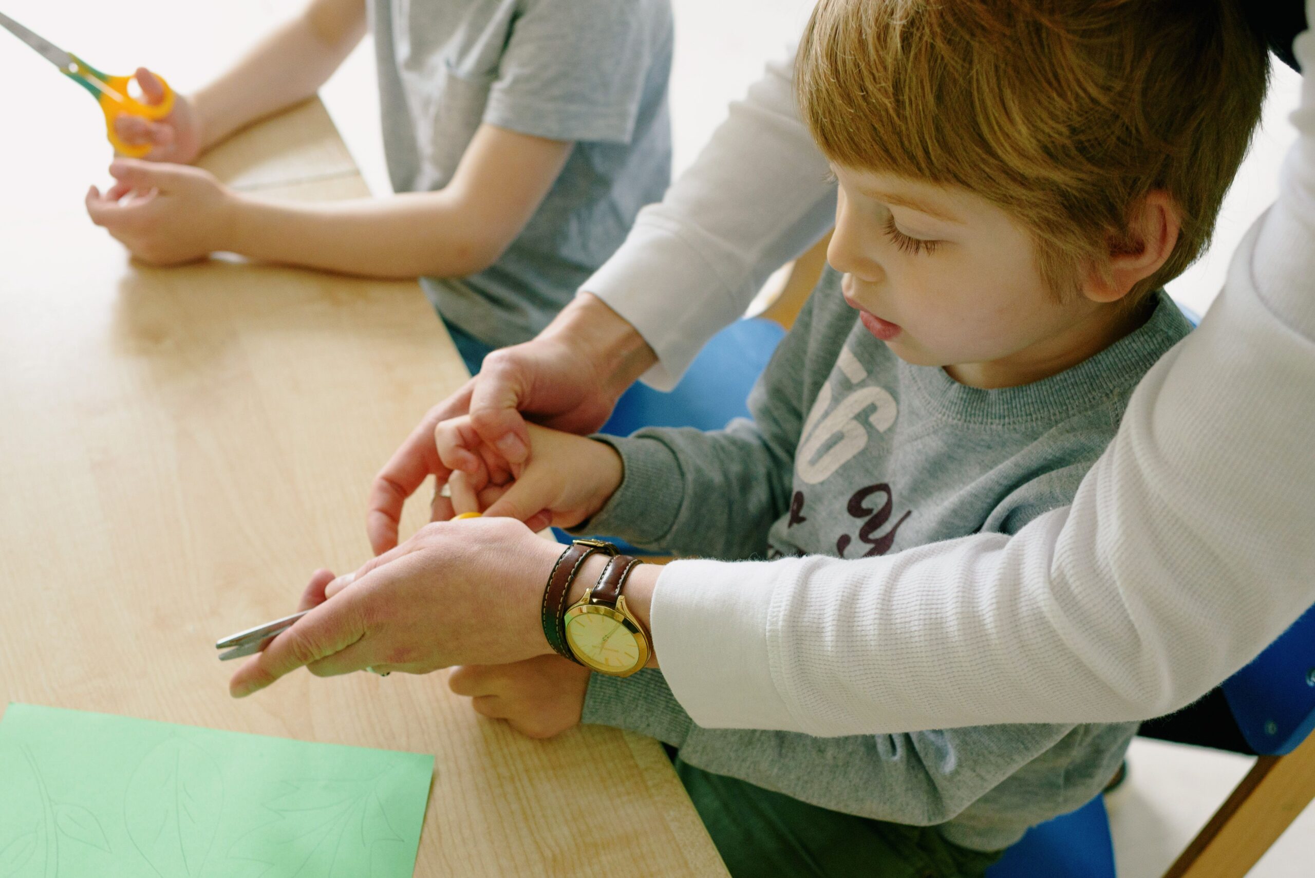 Young child being guided while cutting paper in a school setting. Educational arts activity.